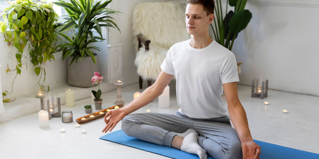 A man in a white t-shirt and gray sweatpants sits cross-legged on a blue yoga mat in a bright, modern room with plants, candles, and a cat in the background.