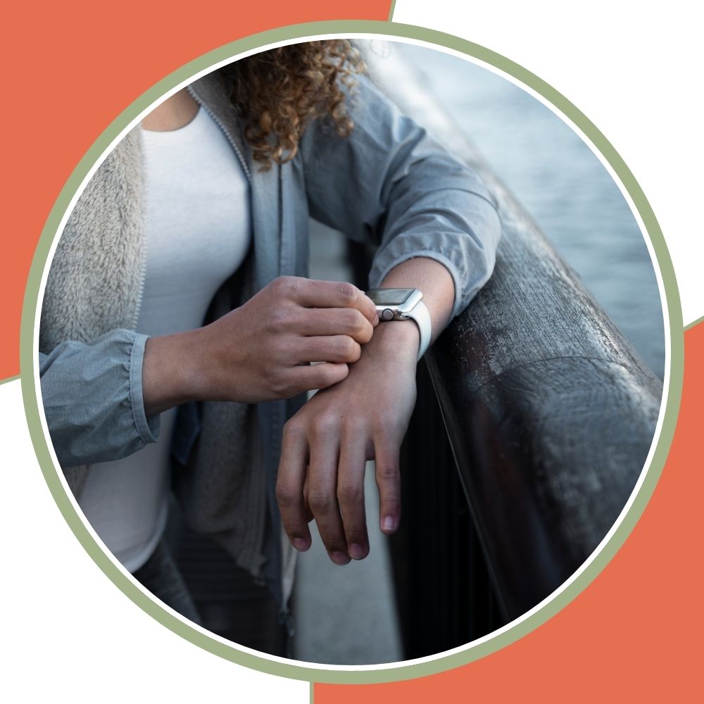 A person in a gray cardigan and white top checks the time on a smartwatch while leaning on a dark railing, framed within a circular border on a coral and white background.